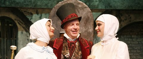 Lavalla kolme näyttelijää, joilla on aikakauden puvut; mies, jolla on korkea musta hattu ja koristeellinen punainen takki, seisoo kahden valkoisiin hattuihin ja mekkoihin pukeutuneen naisen välissä hymyillen maalaistyylisen, maalatun taustan edessä.