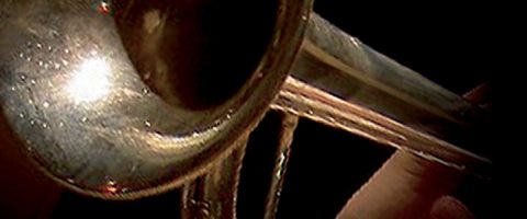 Lähikuva hopeanvärisestä kiiltävästä trumpetista, jota soitetaan, kun sormet painavat venttiilejä tummaa taustaa vasten.