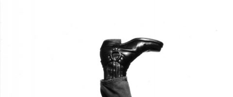 Kiiltävä musta pukukenkä, jossa on metallinen mekaaninen jalka ja nilkka ja jossa on farkut, osoittaa ylöspäin valkoista taustaa vasten.