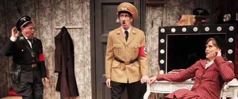 Kolme näyttelijää esiintyy lavalla komediallisessa kohtauksessa; kahdella on liioitellut natsiunivormut, kun taas kolmas, nainen, istuu raidalliseen pukuun pukeutuneena turhamaisen pöydän ääressä ja näyttää tyrmistyneeltä. Lavastuksessa on kukkatapetti ja valaistu peili.