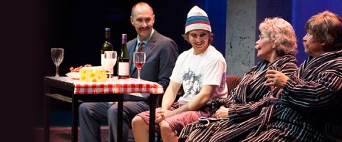 Neljä ihmistä istuu yhdessä pöydän ääressä, jossa on viiniä ja juustoa. Yksi pukumies hymyilee, kun taas toinen, t-paita ja pipo päässä, katsoo kahta vanhempaa naista raidallisissa kaapuissa, jotka kaikki keskustelevat.