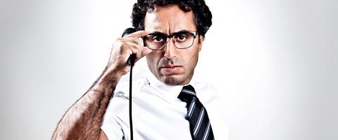 Kiharatukkainen mies, jolla on silmälasit, valkoinen paita ja raidallinen solmio, tuijottaa kameraan intensiivisesti pitäen puhelimen luuria päässään valkoista taustaa vasten.