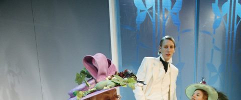 Kolme ihmistä taidokkaissa puvuissa; yhdellä on violetti kukkahattu, toinen valkoisessa puvussa seisoo takana, ja kolmannella on vihreä hattu. He ovat sinisen seinän edessä, jossa on kukkakuvioita.