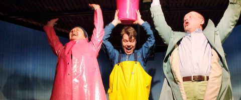 Kolme värikkäisiin sadetakkeihin ja sadevarusteisiin pukeutunutta ihmistä kurottautuu innoissaan ylöspäin, kun keskimmäinen henkilö pitää vaaleanpunaista ämpäriä päänsä yläpuolella, mikä viittaa siihen, että he aikovat pyydystää tai kaataa vettä.