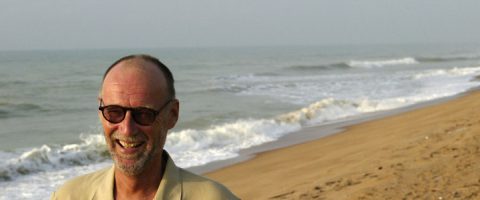 Hymyilevä mies, jolla on silmälasit, seisoo hiekkarannalla, jonka taustalla on lempeät aallot ja pilvinen taivas.