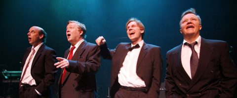 Neljä tummiin pukuihin ja solmioihin pukeutunutta miestä seisoo lavalla sinisessä valaistuksessa ja näyttää laulavan tai esiintyvän yhdessä ilmeikkäillä kasvoilla ja eleillä.