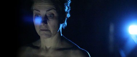 Lyhythiuksinen henkilö valaistaan sinisellä valolla pimeässä ympäristössä. Hän näyttää katsovan alaspäin neutraali ilme kasvoillaan, ja hänen takanaan oikealla näkyy kirkas valonlähde.