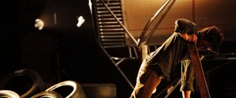 Henkilö kumartuu nostaessaan suurta puulankkua renkaiden ja teollisuuslaitteiden ympäröimänä, ja lämmin valaistus valaisee kohtauksen.