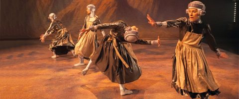 Neljä tanssijaa metallisissa mekoissa ja futuristisissa päähineissä tekee lavalla ilmeikkäitä liikkeitä, kukin ainutlaatuisessa asennossa, lämpimän, pehmeästi valaistun taustan edessä.