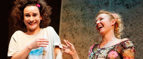 Kaksi naista seisoo yhdessä, molemmat hymyilevät ja nauravat. Toisella naisella on kiharat hiukset, joissa on vaaleanpunainen klipsi, ja hän näyttää hieman yllättyneeltä, kun taas toinen nainen, jolla on vaaleammat hiukset, näyttää leikkisästi kiusoittelevan häntä.