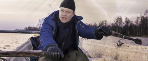 Mustaan pipoon, siniseen takkiin ja hanskoihin pukeutunut henkilö soutaa venettä vesistöllä, jonka taustalla näkyy puita ja silta pilvisen taivaan alla.