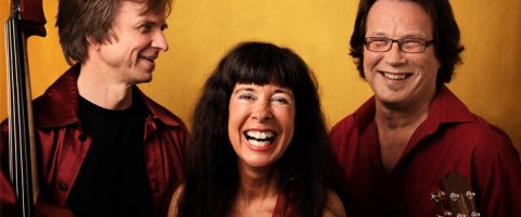 Kolme punapaitaista aikuista seisoo yhdessä hymyillen ja nauraen keltaisen taustan edessä. Yhdellä on kädessään jousisoitin, kun taas keskellä oleva nainen seisoo kahden miehen välissä.