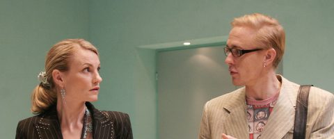 Nainen ja mies seisovat sisätiloissa vastakkain ja keskustelevat. Naisella on taaksepäin vedetyt vaaleat hiukset ja hänellä on puikkopuku, kun taas miehellä on silmälasit ja hänellä on beige takki ja kuviollinen paita.