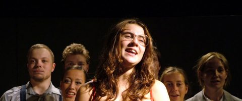 Kuuden nuoren aikuisen ryhmä seisoo tiiviisti yhdessä hämärässä valaistuksessa. Edessä on silmälasipäinen, pitkätukkainen nainen, joka hymyilee ja näyttää laulavan, kun muut katsovat hänen takanaan.