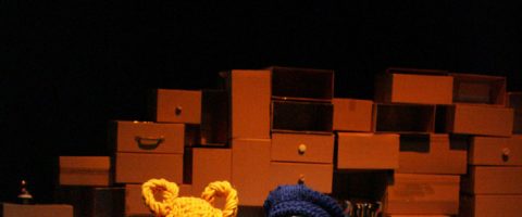 Pahvilaatikkopinoja on kasattu tummaa taustaa vasten. Edessä, kuvan alareunassa, näkyvät kahden kirkkaanvärisen, keltaisen ja sinisen neuletakin päälliset.