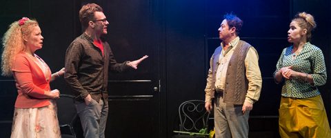 Lavalla neljä näyttelijää keskustelee. Kaksi miestä ja kaksi naista seisovat vastakkain rennoissa ja värikkäissä asuissa, ja taustalla on tumma teatterikulissi.