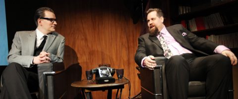 Kaksi pukumiestä istuu nojatuoleissa vastakkain keskustellen. Heidän välissään pienellä pöydällä on vanha kiertävä puhelin. Taustalla näkyy kirjahyllyjä.