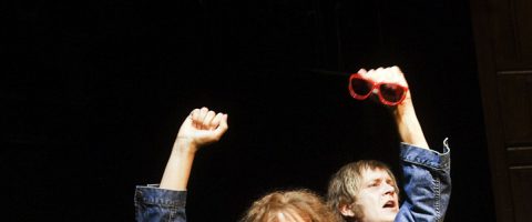 Kaksi sinisiin takkeihin pukeutunutta ihmistä nostaa nyrkkinsä ilmaan; toisella on kädessään punaiset aurinkolasit. Tausta on tumma, ja kohtaus näyttää vangitsevan jännityksen tai juhlan hetken.