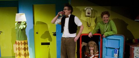 Neljä ihmistä oudossa huoneessa: yksi, jolla on lamppu päänä, yksi juo lasista, yksi vihreässä hymyilee ja neljäs kyyristelee sinisen jääkaapin sisällä. Kirkkaanvärinen lavastus lisää leikkisää tunnelmaa.