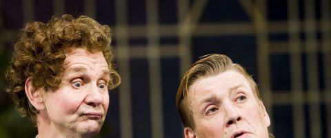 Kaksi ilmeikkään näköistä miestä seisoo lähekkäin, toisella on kihara tukka ja hämmentynyt katse, toisella on liukuhiukset ja koholla oleva kulmakarva, sumean geometrisen taustan edessä.