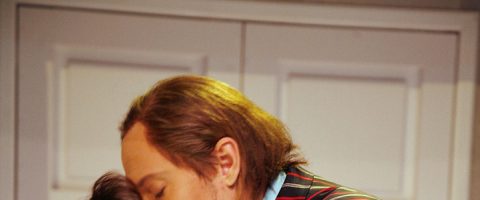 Vaaleanruskeahiuksinen henkilö, jolla on raidallinen takki, nojaa eteenpäin, osittain peitettynä, suorakulmaisilla paneeleilla varustetun valkoisen oven edessä.