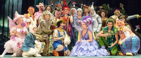 Suuri joukko värikkäisiin, omituisiin pukuihin pukeutuneita ihmisiä, kuten keijuja, eläimiä ja Pinokkion ja Peltimiehen kaltaisia hahmoja, poseeraa yhdessä lavalla metsäaiheisen taustan edessä.