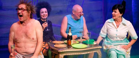 Neljä aikuista istuu pienen pöydän ääressä juomien ja ruoan kanssa nauraen ja keskustellen. Kaksi miestä on ilman paitaa, yhdellä naisella on kirkas mekko, ja toisella on musta asu ja rohkea kampaus. Kohtaus vaikuttaa vilkkaalta ja iloiselta.