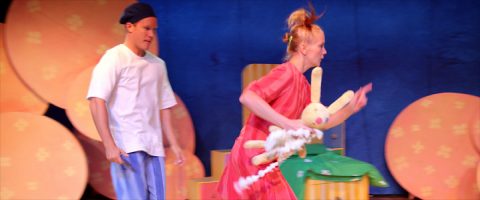 Valkoiseen paitaan ja sinisiin housuihin pukeutunut henkilö seisoo punamekkoon pukeutuneen henkilön takana, jolla on kädessään suuri pehmojänis. Molemmat näyttävät olevan lavalla, jonka taustalla on suuria oransseja ympyröitä.