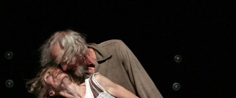 Iäkäs mies ja nainen tanssivat lavalla tiiviisti yhdessä, ja mies nojaa hymyilevän naisen päälle. Molemmat vaikuttavat iloisilta. Taustalla on pimeää, ja näkyvissä on pieniä pyöreitä valoja.