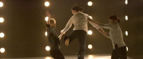 Kolme ihmistä esittää nykytanssia lavalla, ja yksi hyppii, kun kaksi muuta pitävät häntä kädestä. Pyöreät näyttämövalot muodostavat taustalla rivejä, jotka luovat dramaattisen, valaistun ympäristön.