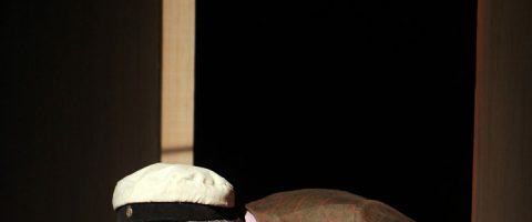 Henkilö, jolla on valkoinen lippis ja ruskea ruudullinen takki, on kumartuneena tumman taustan edessä, ja osa hänen vartalostaan on valaistu.