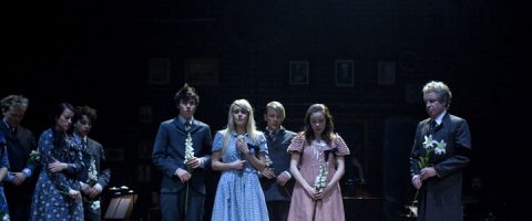 Joukko ihmisiä seisoo hämärästi valaistulla näyttämöllä valkoisia kukkia kädessään. Useimmilla on synkät ilmeet ja vanhanaikaiset vaatteet, ja keskellä on kaksi kuviomekkoon pukeutunutta tyttöä. Tausta on tumma, ja seinällä on kehystettyjä kuvia.