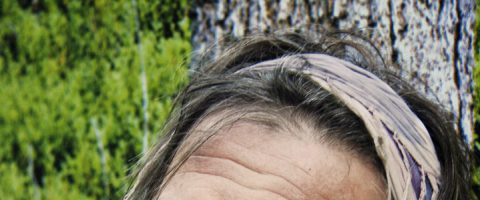 Lähikuva henkilön pään yläosasta, jolla on vaalea päähine, taustalla on puun kuorta ja vihreitä lehtiä.