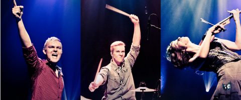 Kolme muusikkoa esiintyy energisesti lavalla dramaattisessa valaistuksessa, kaksi soittaa rumpuja ja yksi viulua, ja jokainen heistä on kuvattu ilmeikkäissä ja dynaamisissa asennoissa.