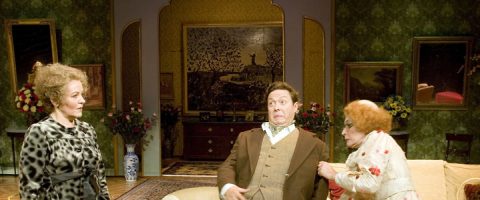 Kolme aikakauden pukuihin pukeutunutta näyttelijää esiintyy koristeellisella lavasteella, vasemmalla yksi nainen kuvioidussa takissa, keskellä mies, joka näyttää yllättyneeltä, ja toinen kukkamekkoon pukeutunut nainen, joka vetää miestä kädestä.