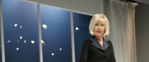 Lyhyet vaaleat hiukset omaava nainen, jolla on tumma puku, seisoo sisätiloissa suurten ikkunoiden edessä, joissa on siniset paneelit ja valkoinen verho.