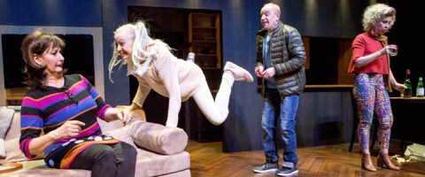 Neljä aikuista on olohuoneessa. Yksi nainen istuu sohvalla säikähtäen, kun toinen nainen hyppää leikkisästi häntä kohti. Mies seisoo lähellä, ja värikkäisiin housuihin pukeutunut nainen valmistelee juomia pöydässä taustalla.