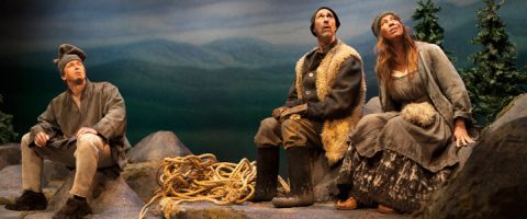 Kolme maalaismaisesti vanhanaikaisiin vaatteisiin pukeutunutta näyttelijää istuu näyttämöllä kivillä, mäntyjen ja köyden ympäröimänä, ja katselee ylöspäin vakavin ilmein maalattua vuoristotaustaa vasten.