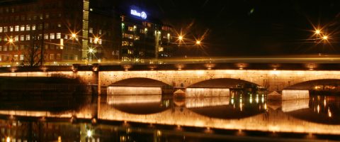 Yöllä katuvalojen valaisema kivisilta heijastuu alla olevaan rauhalliseen jokeen, ja läheiset rakennukset ovat myös valaistuja, mikä luo rauhallisen yöllisen kaupunkimaiseman.