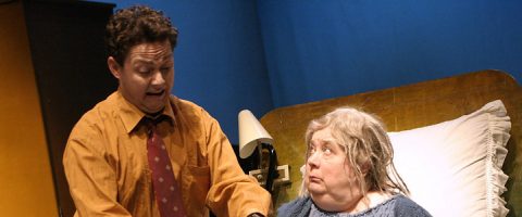 Oranssiin paitaan ja punaiseen solmioon pukeutunut mies seisoo vanhemman harmaahiuksisen naisen vieressä, joka istuu sängyssä ja katsoo häntä hämmästyneenä. Heidän takanaan pöydällä on lamppu.