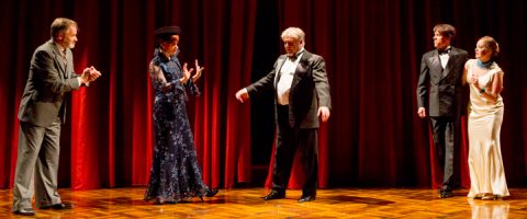 Viisi näyttelijää esiintyy dramaattisesti näyttämöllä, jossa on punaiset verhot ja puulattia. Kohtaus on ilmaisuvoimainen, ja siinä on monipuolisia eleitä ja intensiivistä sitoutumista hahmojen välillä.