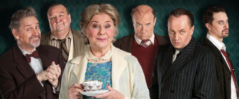 Kuusi vintage-vaatteisiin pukeutunutta aikuista poseeraa yhdessä; keskellä oleva vanhempi nainen pitelee teekuppia, ja häntä ympäröi viisi miestä, joilla on vaihtelevat ilmeet, ja kaikki seisovat tummanvihreää taustaa vasten.