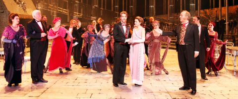 Suuri joukko tyylikkäisiin, 1900-luvun alun pukuihin pukeutuneita näyttelijöitä seisoo kirkkaasti valaistulla näyttämöllä hymyillen ja elehtien kuin musikaali- tai teatteriesityksen aikana. Lavastuksessa on upea, vintage-henkinen tausta.