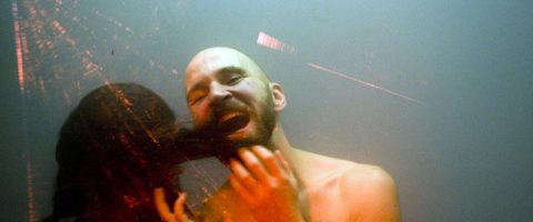 Kalju, parrakas mies seisoo ilman paitaa lasin takana hymyillen suu auki, kun hänen viereensä ilmestyy tumma, epäselvä hahmo, joka luo surrealistisen ja levottoman vaikutelman.