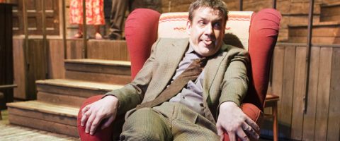 Ruudulliseen pukuun pukeutunut mies istuu punaisessa nojatuolissa ja työntää leikkisästi kieltään ulos. Taustalla lavalla on puisia portaita ja toinen henkilö, jolla on kukkahousut.