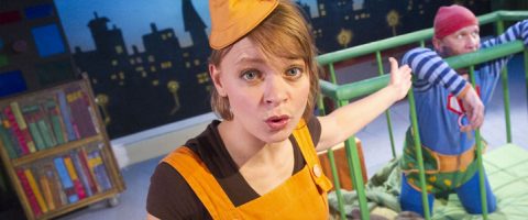 Oranssiin asuun ja hattuun pukeutunut nainen elehtii yllättyneenä vihreän pinnasängyn vieressä, kun taas värikkäisiin vaatteisiin ja punaiseen pipoon pukeutunut mies istuu pinnasängyn sisällä. Heidän takanaan näkyy maalattu kaupunkitausta ja kirjahylly.