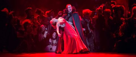 Dramaattinen näyttämökohtaus, jossa mustaan viittaan pukeutunut mies pitelee punaiseen mekkoon pukeutunutta naista, joka nojaa taaksepäin. Heitä ympäröi tummiin pukuihin pukeutunut väkijoukko punaisessa valaistuksessa, mikä luo goottilaisen, teatterimaisen tunnelman.