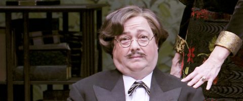 Aaltoileva tukka, pyöreät silmälasit ja viikset omaava mies pukeutuu smokkiin, jossa on rusetti. Hän istuu kirjahyllyjen edessä, kun naisen käsi lepää hänen olkapäällään.