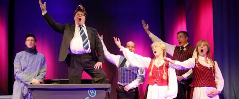 Pukuun ja kruunuun pukeutunut mies seisoo pöydän päällä käsi koholla, ja häntä ympäröi viisi värikkäisiin pukuihin pukeutunutta ihmistä, jotka kaikki laulavat tai huutavat dramaattisin ilmein violetilla ja vaaleanpunaisella värillä valaistulla näyttämöllä.