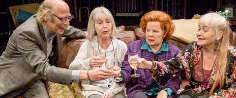 Neljä iäkästä aikuista istuu yhdessä sohvalla, hymyilee ja kohottaa lasit maljan kohotukseksi. Seurue vaikuttaa iloiselta ja juhlalliselta.